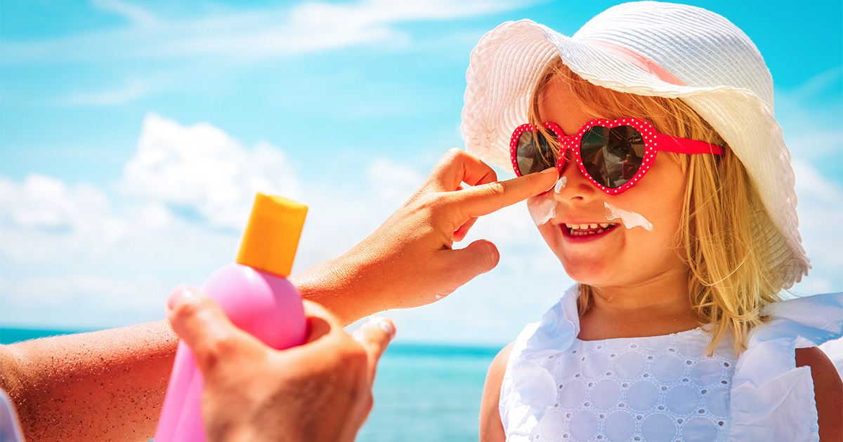 A young girl wearing a sun hat and heart-shaped sunglasses has sunscreen applied on her face at a beach.