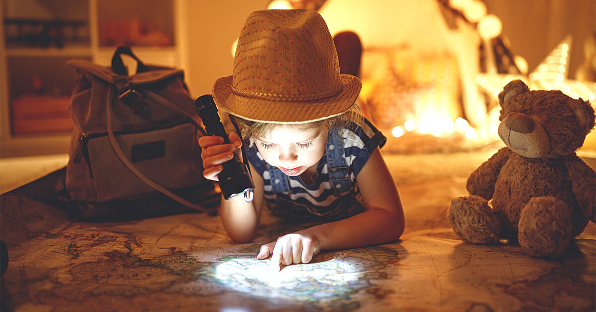 Child using a flashlight to explore a map with a teddy bear and backpack nearby.